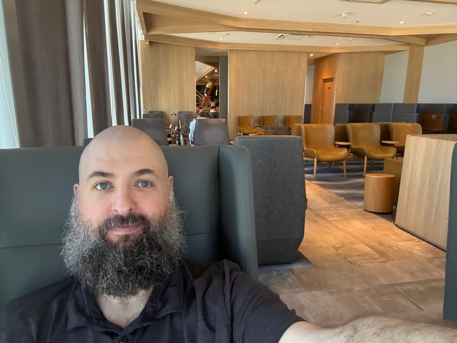 Selfie in the Plaza Premium Lounge, Mactan-Cebu International Airport, bald man with a full dark beard in a black shirt, in a high-backed grey lounge chair, warm wood paneling and amber leather seating behind him